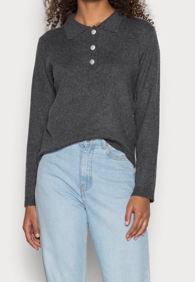 Pull en tricot gris avec un col, ornée de trois boutons décoratifs et de manches longues, assorti à un jean taille haute bleu clair.