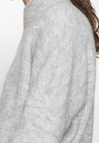 Světle šedý pletený svetr s vysokým límcem, který má žebrovanou texturu a jemný diagonální vzor na rukávech.