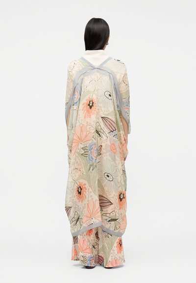 Γυναίκα με μακριά σκουρόχρωμα μαλλιά φοράει μια ιριδίζουσα, διάφανη κάπα με floral σχέδιο και αντίστοιχο παντελόνι με φαρδιά πόδια, στέκεται μπροστά σε απλό φόντο.