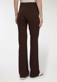 Osoba nosząca ciemnobrązowe, wysokie, szerokie spodnie stoi z rękami luźno wzdłuż ciała, w połączeniu z białymi butami i jasną górą.