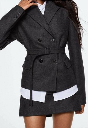 Femme portant un blazer croisé gris foncé ceinturé sur une chemise blanche, assorti à une jupe gris foncé, debout devant un fond blanc.