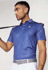 Polo shirt blu navy con motivo a rombi, maniche corte e colletto. Presenta un piccolo logo sul petto. Abbinato a una cintura grigia e un bastone da golf.
