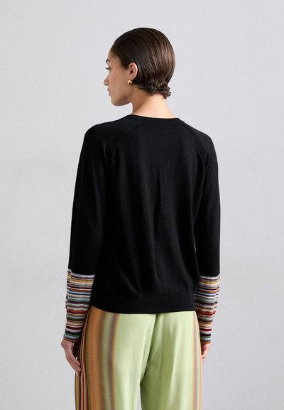 Maglione nero con maniche a strisce multicolori, caratterizzato da un collo rotondo e design raglan. Il modello indossa pantaloni a righe verde chiaro.