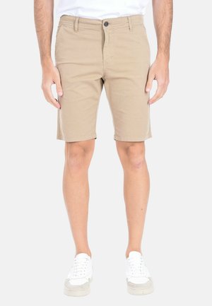 Uomo che indossa pantaloncini beige al ginocchio e sneakers bianche in piedi davanti a uno sfondo semplice.