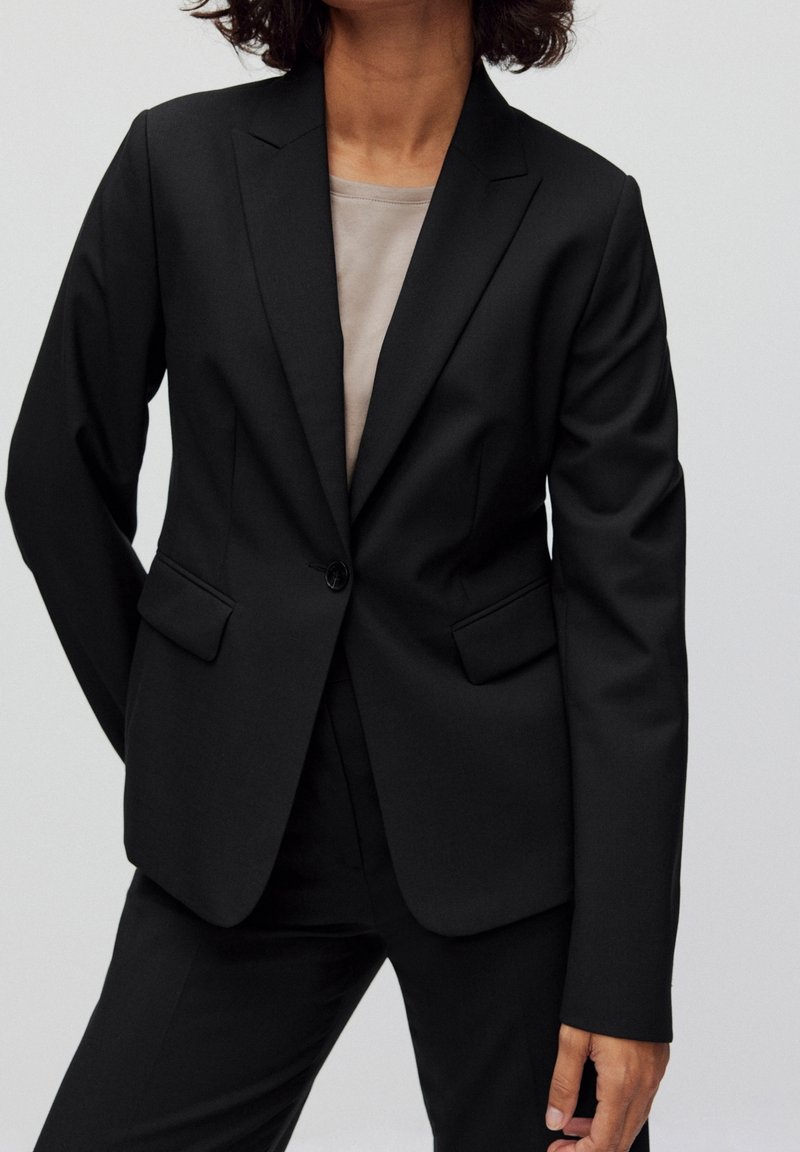 Personne portant un blazer noir sur mesure à un seul bouton par-dessus un haut beige avec un pantalon noir assorti, sur un fond uni.