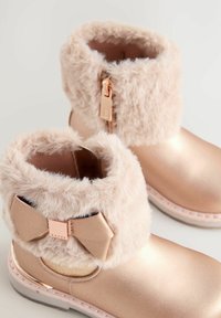 Botines de oro rosa con un puño de pelo suave, cremallera lateral y un pequeño detalle de lazo. Exteriores lisos con acentos de costura.