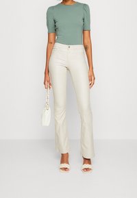 Mörkgrön topp med korta puffärmar i passform, tillsammans med ljusbeige utsvängda byxor och krämfärgade flätade sandaler; håller en vit handväska.