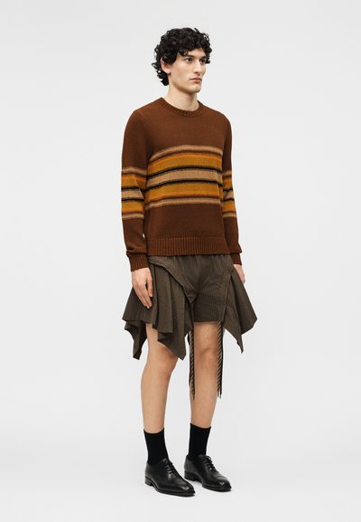 Un mannequin masculin porte un pull marron à rayures, une jupe marron asymétrique avec un tissu drapé, des chaussettes noires et des chaussures habillées noires sur un fond uni.