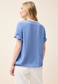 Blusa azzurro chiaro a maniche corte con risvolti, scollatura rotonda e fondo arricciato. Realizzata in un tessuto liscio, abbinata a pantaloni bianchi.
