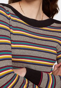 Mehrfarbiger gestreifter Pullover mit einem schwarzen Rippkragen. Er hat horizontale Streifen in Rot, Gelb, Grün und Blau auf einem neutralen Hintergrund.