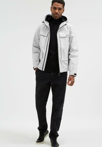Chaqueta blanca impermeable con capucha, dos bolsillos frontales y puños ajustables, llevada sobre un suéter negro de cuello alto y combinada con pantalones y zapatos negros.