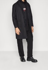 Cappotto in misto lana nero con revers, tasche frontali e chiusura a bottoni, indossato sopra un maglione nero e abbinato a pantaloni e scarpe nere.