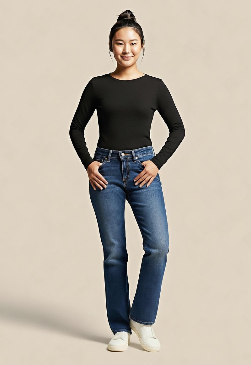 Giovane donna in piedi con le mani nelle tasche dei jeans, indossando un top nero a maniche lunghe, jeans blu, scarpe bianche e un'acconciatura a bun su uno sfondo beige.