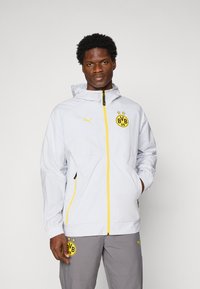 Homme portant une veste Puma Borussia Dortmund blanche avec une fermeture éclair jaune et un pantalon gris, se tenant devant un fond blanc uni.