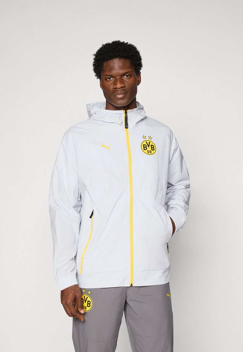 Homme portant une veste Puma Borussia Dortmund blanche avec une fermeture éclair jaune et un pantalon gris, se tenant devant un fond blanc uni.