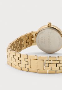 Ρολόι χειρός με μπρασελέ από χρυσόμεταλλο με κούμπωμα, που δείχνει την πίσω πλευρά του στρογγυλού καντράν με χαραγμένο το λογότυπο Calvin Klein και σημάδια.