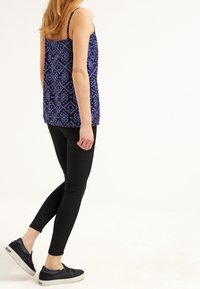 Blå mönstrad linne med tunna axelband, i kombination med svarta åtsittande leggings och mörka slip-on skor, visad från en sidovinkel.