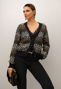 Frau in gemusterter Strickjacke, mit einem mit Spitze verzierten Oberteil, schwarzen Hosen, die eine schwarze Clutch hält und auffällige Ohrringe trägt, posiert vor einem schlichten Hintergrund.