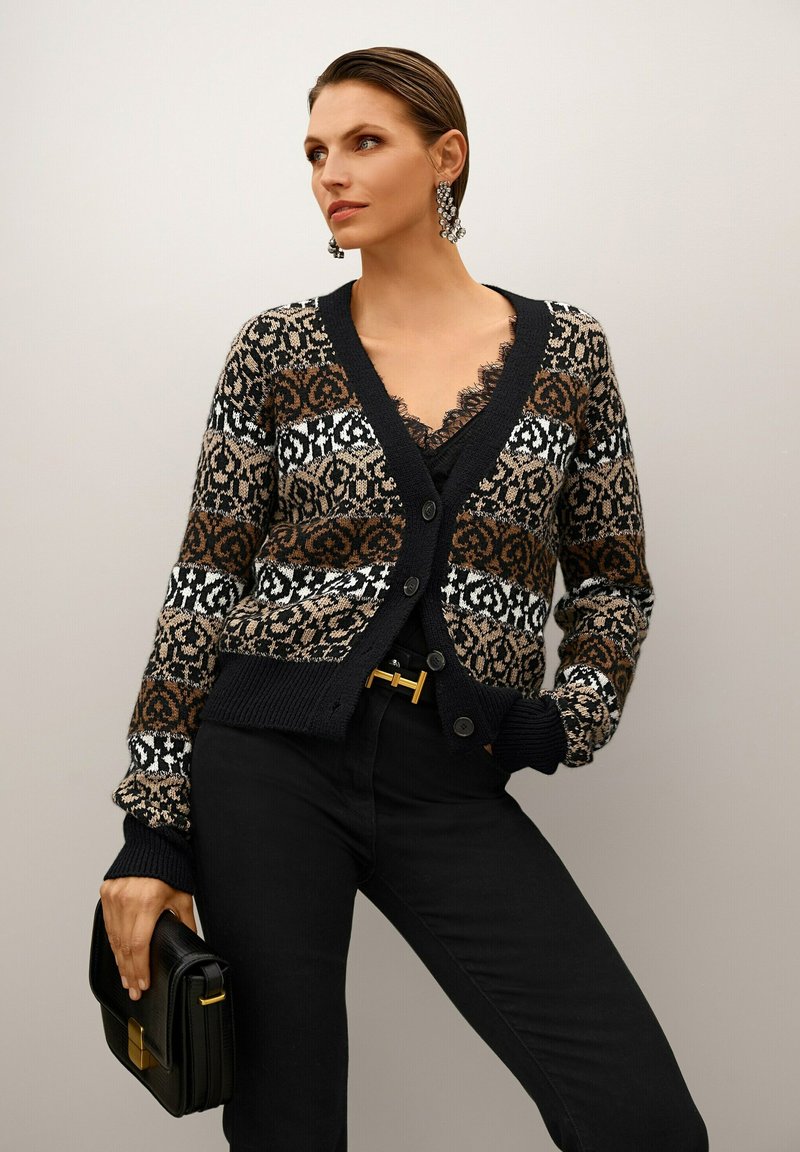 Frau in gemusterter Strickjacke, mit einem mit Spitze verzierten Oberteil, schwarzen Hosen, die eine schwarze Clutch hält und auffällige Ohrringe trägt, posiert vor einem schlichten Hintergrund.