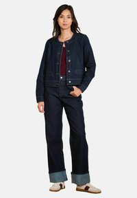 Veste en denim foncé avec un col rond et des boutons sur le devant, associée à un pantalon en denim foncé à jambes larges et des baskets crème avec des accents bruns.
