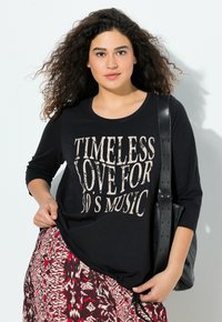 Černé tričko s dlouhým rukávem s potištěným textem "NADČASOVÁ LÁSKA K HUDBĚ 90. LET." V kombinaci se vzorovanou sukní a černou taškou.