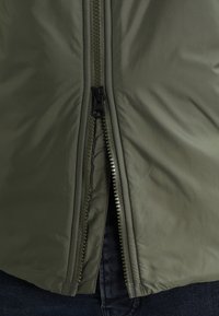 Olivgrön zip-up jacka med en slät yta, kontrasterande svart dragkedja och synlig sömnad längs sömmarna.