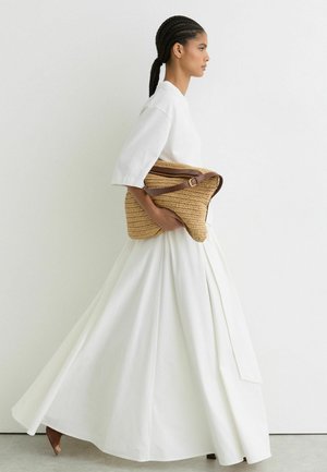 Fonott hajú nő fehér ruhában, nagy, barna bőr pántos világosbarna fonott táskát tartva, fehér háttér előtt sétálva.