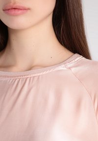 Vaaleanpunainen satiininen lyhythihainen paita, jossa on pyöreä pääntie ja hienovarainen ommelkaulus ja hihoissa. Sujuva pinta.