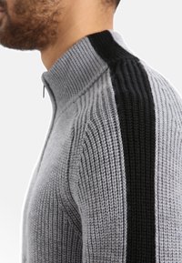 Grauer gestrickter Pullover mit Kragen und Reißverschluss, der auf der linken Seite einen schwarzen senkrechten Streifen und eine gerippte Textur aufweist.