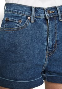 Pantalones cortos de mezclilla en azul medio con un diseño clásico, que incluye detalles de costuras, bolsillos delanteros y traseros, y un dobladillo enrollado.