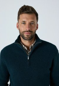 Pulover cu fermoar la jumătate, de culoare verde închis, cu o suprafață texturată, purtat peste o cămașă în carouri. Fermoarul are o tabulator metalic.