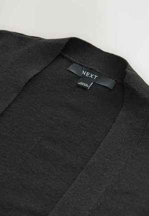 Gilet noir en tissu tricoté. Il présente un col en V et un design à l'avant ouvert, avec une étiquette de marque "NEXT" près du cou.