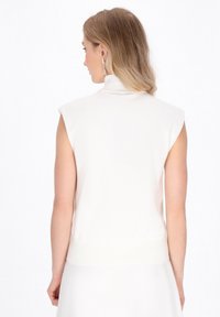 Ærmeløs hvid turtleneck-top, tætsiddende strikket materiale, ribbet tekstur ved kanten og skuldersømmen. Båret af en person, der vender væk.