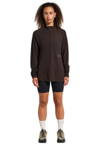 Brun långärmad topp med halvdragkedja, matchad med svarta shorts. Atletiska skor och vita strumpor synliga, med en logotypdetalj.