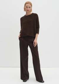 Brauner Strickpullover mit strukturierter Oberfläche, lässigem Schnitt und rundem Halsausschnitt, kombiniert mit hoch taillierten, weit geschnittenen braunen Hosen und dunklem Schuhwerk.