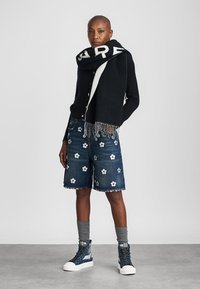 Svart skjerf med hvit skrift, svart ribbestrikket genser, denimshorts med blomsterapplikasjoner, og blå high-top sneakers med teksturerte såler.