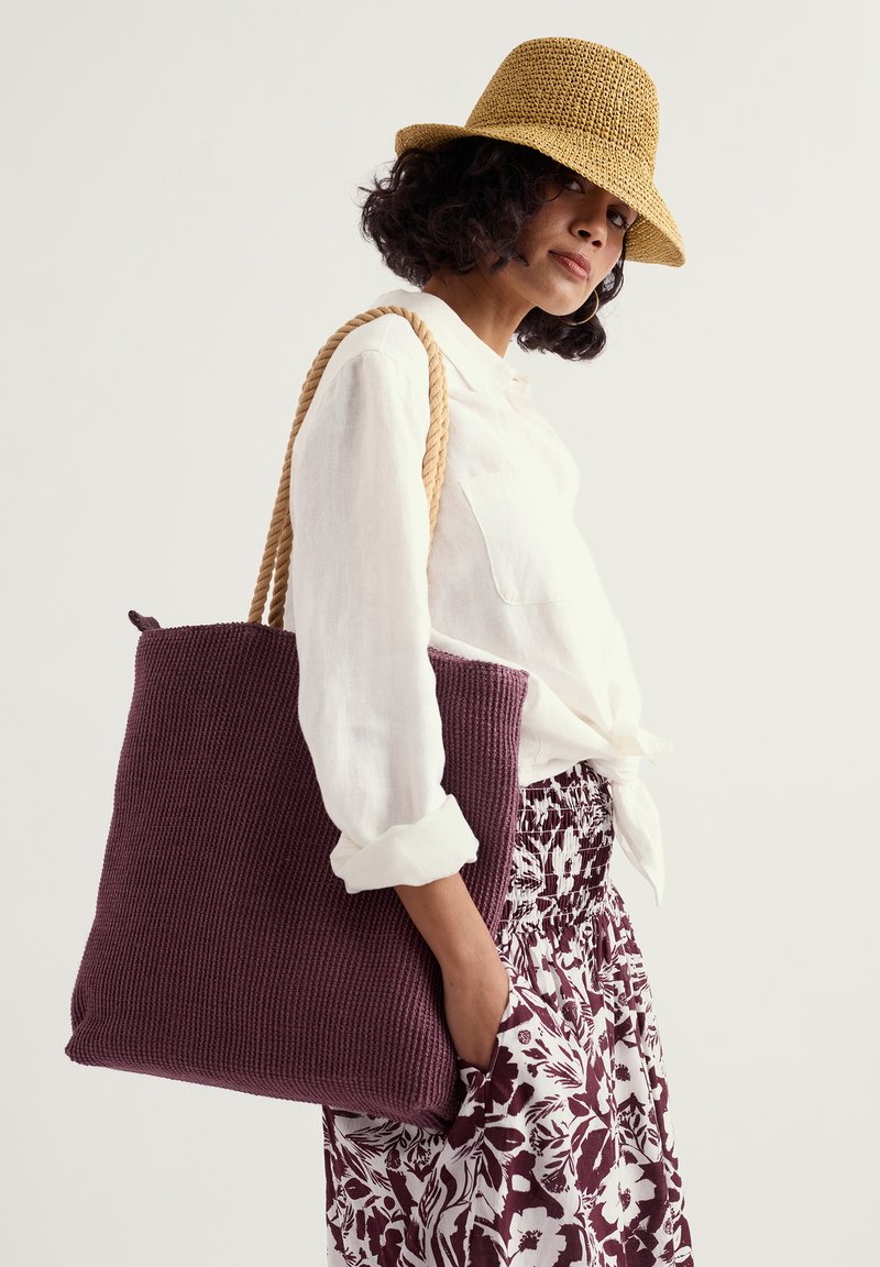 Femme portant un chapeau de paille, une chemise blanche boutonnée, une jupe fleurie, et portant un grand sac cabas violet tricoté avec des poignées épaisses en corde.
