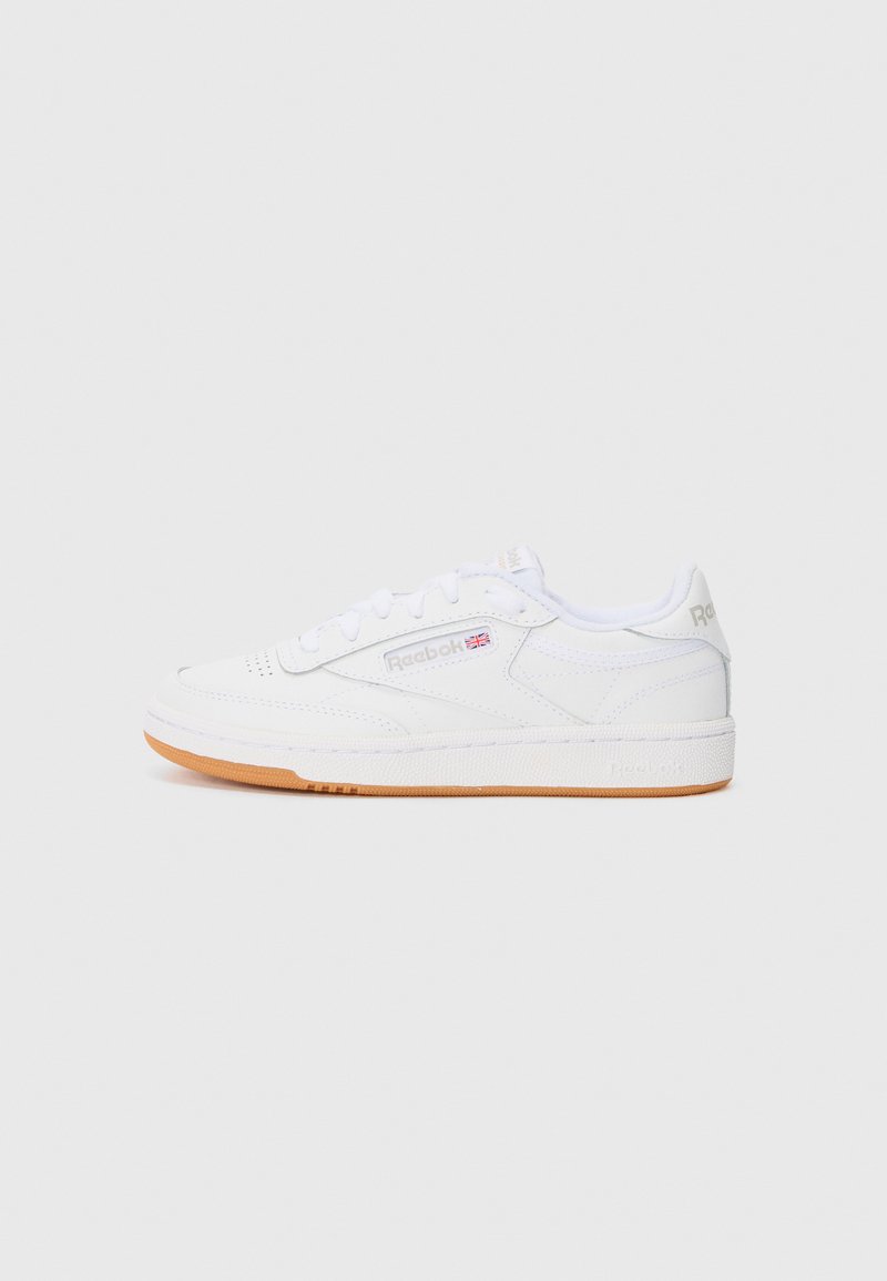 Bílé kožené tenisky s nízkým střihem, které mají texturovanou bílou gumovou podrážku a hnědou gumovou podešev. Přítomné jemné akcenty loga.