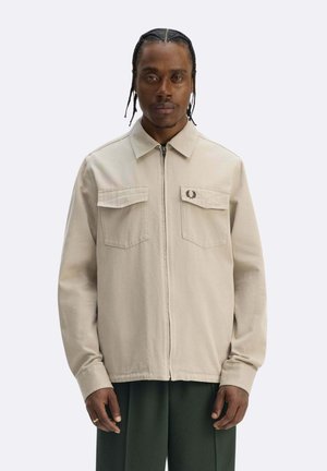 Uomo con capelli intrecciati che indossa una giacca beige con zip e due tasche sul petto, pantaloni verde scuro, in piedi davanti a uno sfondo semplice.