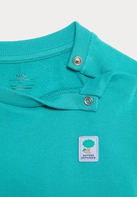Camicetta di cotone turchese con colletto a girocollo a coste, dettaglio di chiusura a bottoni e una toppa rettangolare "Esploratore della Natura" con un design ad albero.