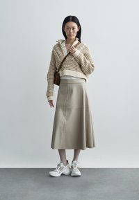Pleteni bež skrajšani pulover, bež usnjena midi krilo, bele superge z zlatimi poudarki. Model stoji pred nevtralnim ozadjem.