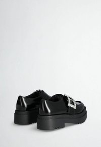 Black patent leather shoes with a chunky black sole, featuring a metallic decorative buckle and a textured upper.