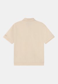 Beżowa koszulka polo z krótkim rękawem i kołnierzykiem, widziana od tyłu, z małym logo na lewym rękawie.