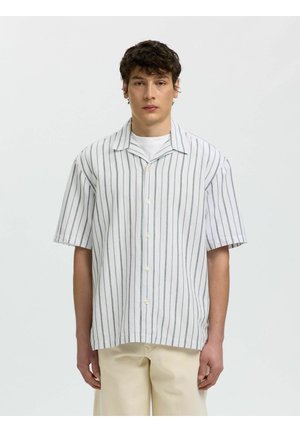 Ung mand iført en hvid kortærmet skjorte med lodrette grå striber og beige bukser, stående mod en ensfarvet lys baggrund.