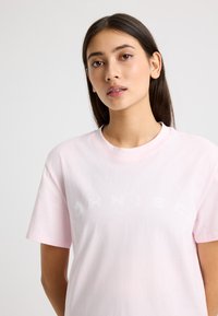 Camiseta de algodón rosa claro con mangas cortas y cuello redondo. Presenta un logo de texto impreso en blanco en la parte delantera. Corte estándar y relajado.