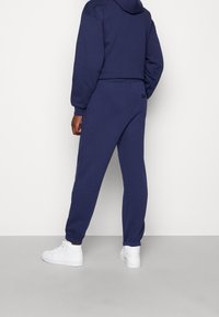 Marinblå hoodie och joggers i mjukt tyg, med elastiska ärmslut och midjeband. Bärs med vita sneakers, bakvy visas.