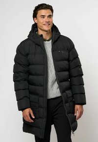 Svart puffjakke med hette, med glidelås foran, ribbede mansjetter og quiltet design. Bærers over en grå genser og bukser.