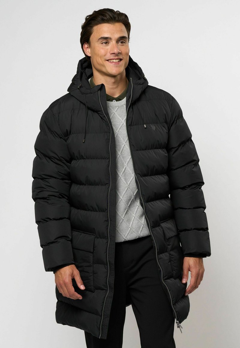 Svart puffjakke med hette, med glidelås foran, ribbede mansjetter og quiltet design. Bærers over en grå genser og bukser.