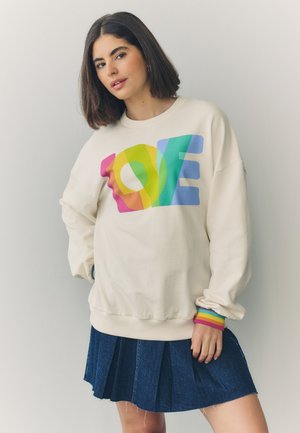 Frau mit schulterlangem dunklem Haar, die einen cremefarbenen Pullover mit buntem "LOVE"-Design und einem blauen Plisseerock vor einfarbigem Hintergrund trägt.