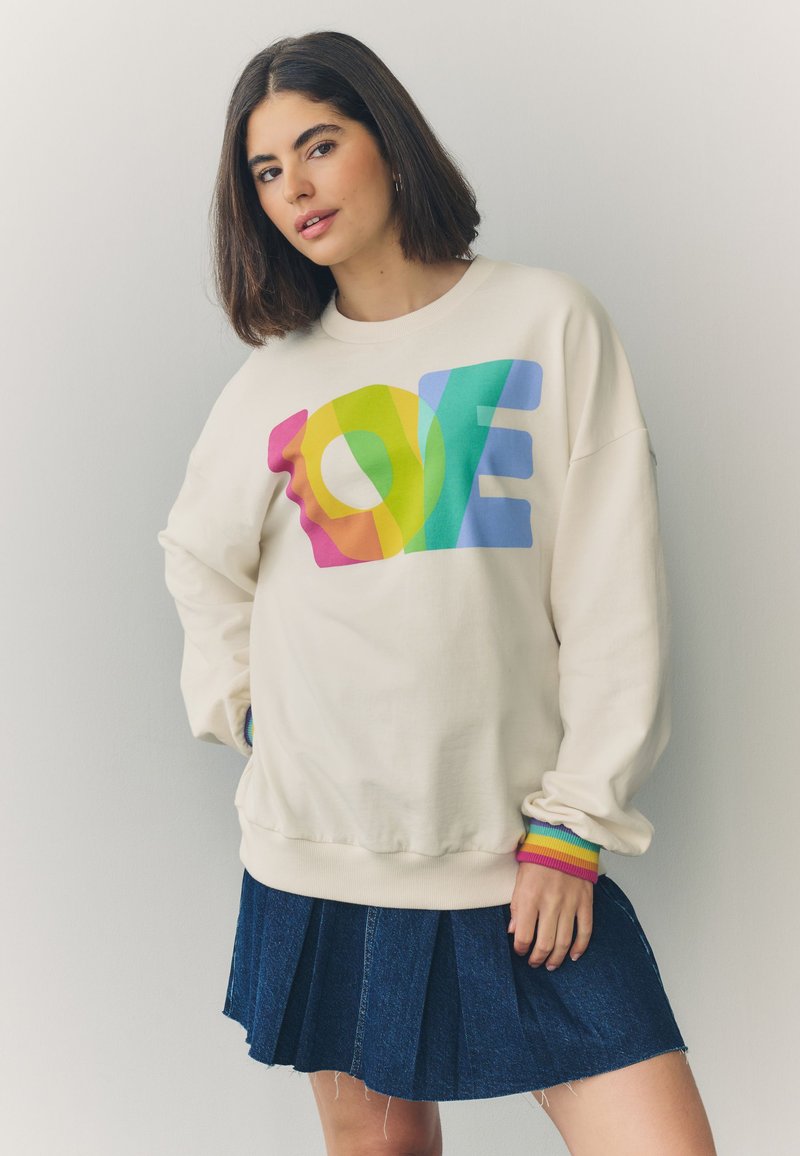 Femme aux cheveux mi-longs et foncés portant un sweat crème avec un motif coloré "LOVE" et une jupe plissée bleue sur un fond uni.